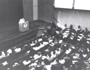 lecturer speaks in front of a large crowd in an auditorium.