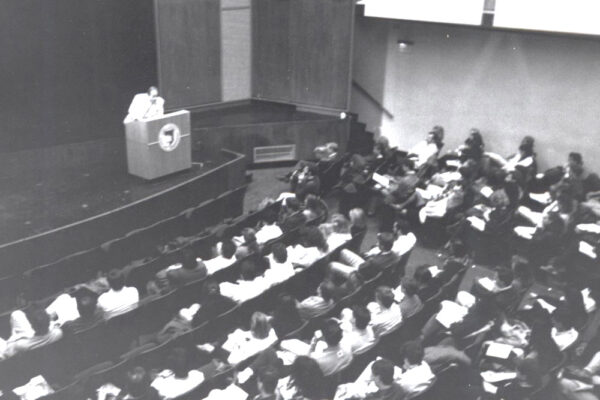 lecturer speaks in front of a large crowd in an auditorium.