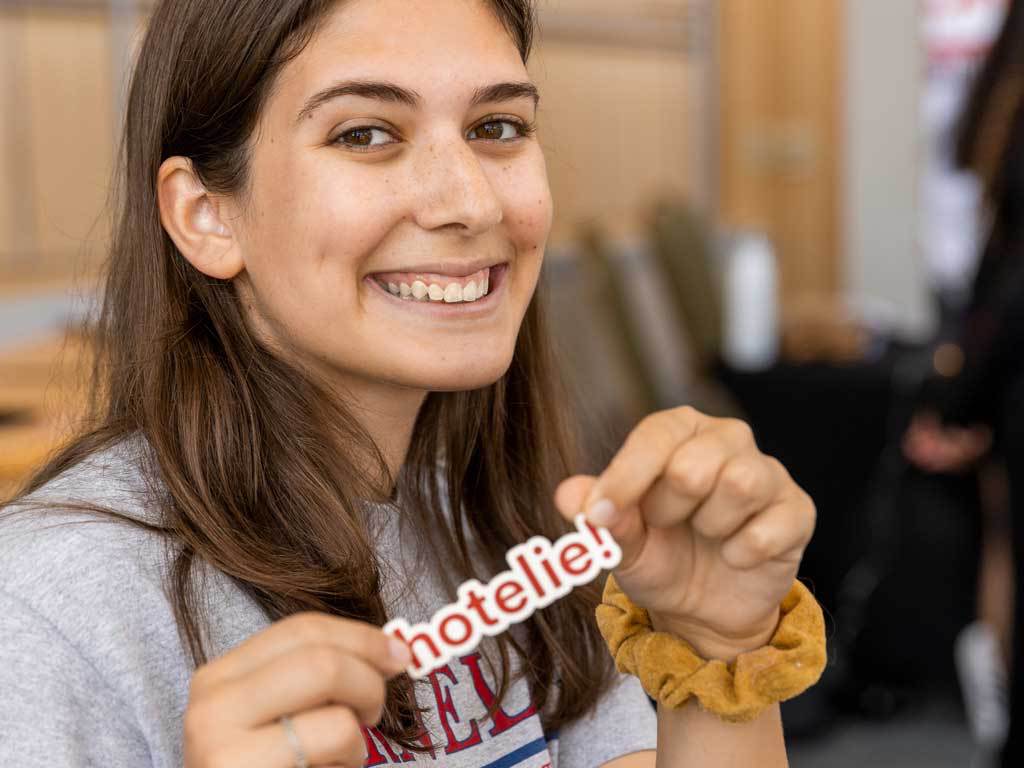 A student with medium-length brown hair smiles holding a sticker that says “hotelie!”