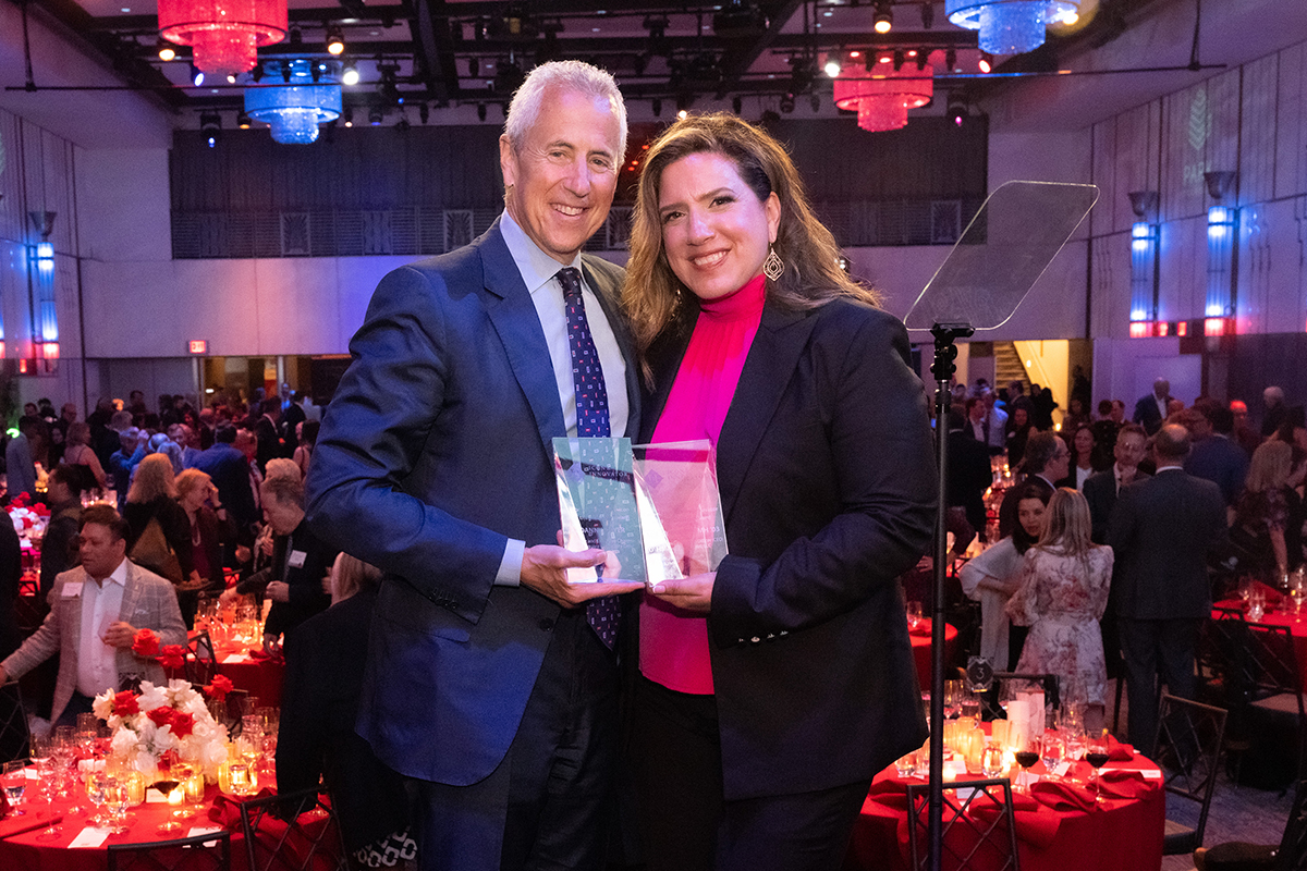 Award-winners Mia Kyricos, MMH ‘03, and Danny Meyer stand on stage at the 14th annual Icons and Innovators Award ceremony, with the crowd and tables behind them.