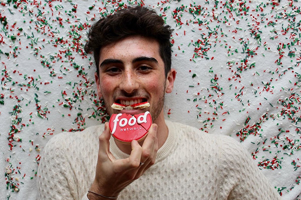 A young man smiling and taking a bite out of a frosted cookie with the word "food" on it.