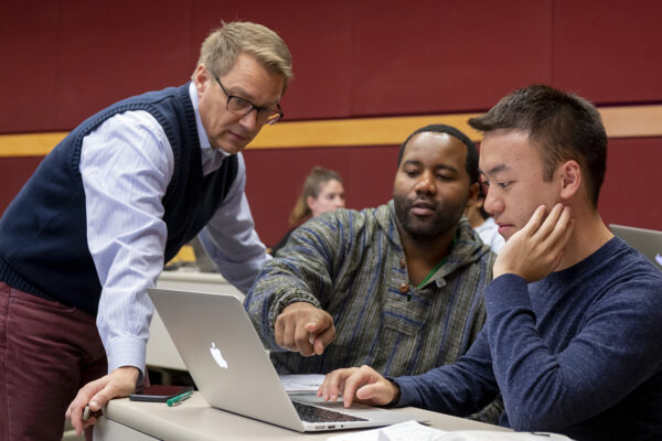 A professor stands looking at a laptop with two seated students.