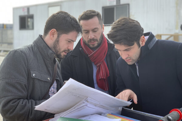 Three men in outerwear stand outside looking at papers.