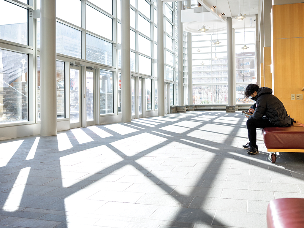 A student sits on a padded bench facing a wall of windows with natural light streaming in.