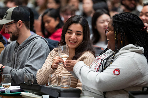 Students sitting next to each other in a crowded lecture hall smile and clink wine glasses before tasting the white wine in them.
