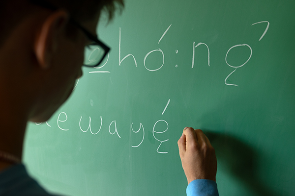 Person writing on a green chalkboard in the Gayogohó:nǫˀ language.