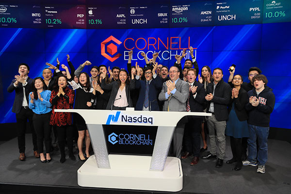 Group photo of Cornell Blockchain members cheering and standing behind the Nasdaq podium at the opening bell, with Cornell Blockchain on the wall behind them and on the podium in front of them