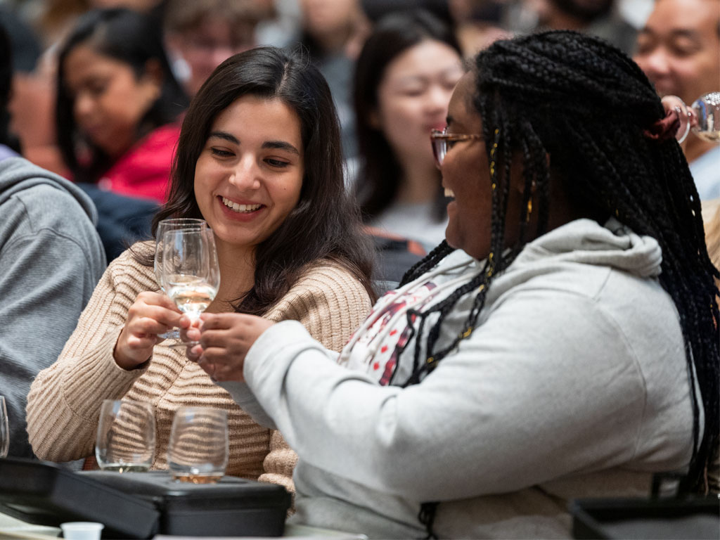 Students sitting in a tiered classroom click their wine glasses in a “cheers.”