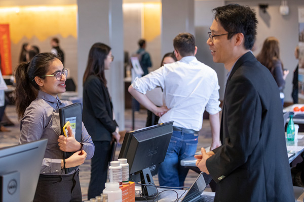 Two people speak over a desktop computer with other small groups of people talking in the background.