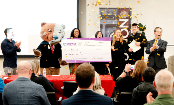 Students stand with a bear mascot and an oversized check in front of an audience as gold confetti falls.