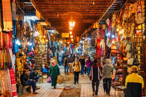 A bustling market street lined with colorful shops and stalls displays textiles, bags, and lanterns, with people walking through under a lattice-covered ceiling illuminated by hanging lanterns.