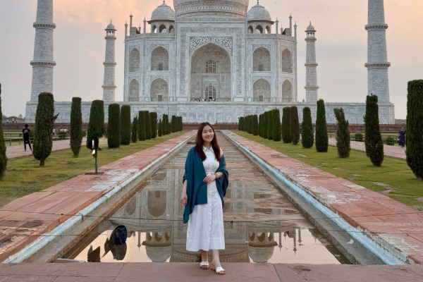 Vanessa stands in front of the Taj Mahal, which is reflected in a long rectangular pool beneath a sky dotted with clouds.