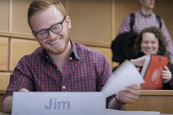 Person named Jim smiling and flipping through notes in a lecture hall setting.