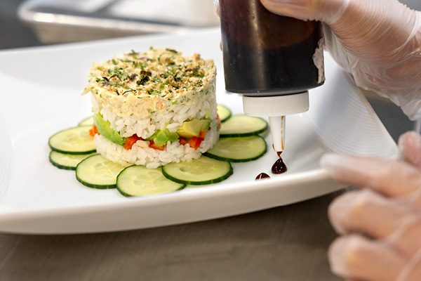 A gloved hand squeezes dots of sauce around a dish of rice and vegetables.