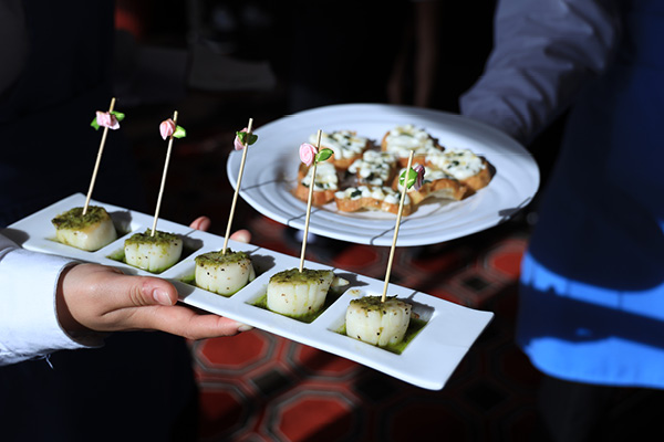 A close-up of two people carrying small portions of scallops and crostini.